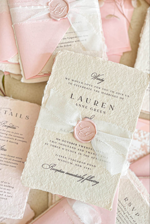 This handmade paper wedding invitation suite exudes timeless elegance with a touch of modern artistry. Crafted on luxury cotton paper with deckled edges, each invitation tells a story of care and craftsmanship. The delicate texture pairs beautifully with blush and ivory tones, creating a look that feels both classic and ethereal. Every detail is completed with a wax seal monogram, adding a touch of old-world refinement.