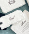 Close-up of a personalized ivory handmade paper wedding place card featuring the name Val in elegant black calligraphy and naturally deckled edges. The card rests over a coordinating handmade paper wedding menu with black script typography and a custom floral crest monogram, styled on textured dusty blue fabric with black chiffon ribbon.