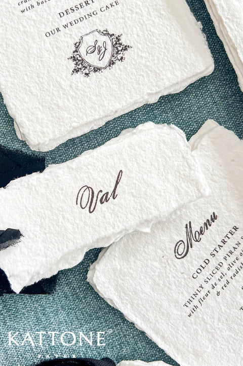 Close-up of a personalized ivory handmade paper wedding place card featuring the name Val in elegant black calligraphy and naturally deckled edges. The card rests over a coordinating handmade paper wedding menu with black script typography and a custom floral crest monogram, styled on textured dusty blue fabric with black chiffon ribbon.