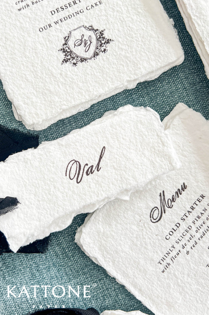 Close-up of a personalized ivory handmade paper wedding place card featuring the name Val in elegant black calligraphy and naturally deckled edges. The card rests over a coordinating handmade paper wedding menu with black script typography and a custom floral crest monogram, styled on textured dusty blue fabric with black chiffon ribbon.