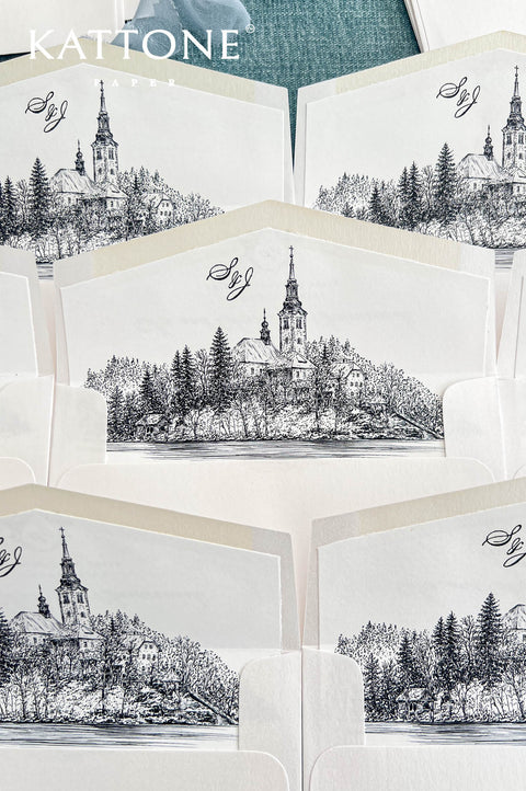 Close-up of an elegant ivory wedding envelope featuring a custom black ink illustration of a historic lakeside church in Lake Bled, Slovenia, surrounded by detailed trees and waterfront scenery. Personalized calligraphy monogram above the illustrated liner, paired with a handmade paper wedding invitation featuring a black floral crest, black silk ribbon and white monogram wax seal.