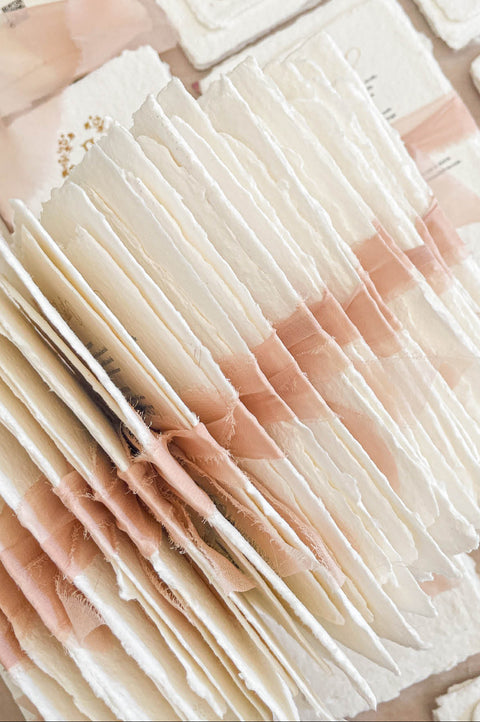 Stacked handmade paper wedding invitations and enclosure cards showcasing custom monogram crest and soft blush accents.