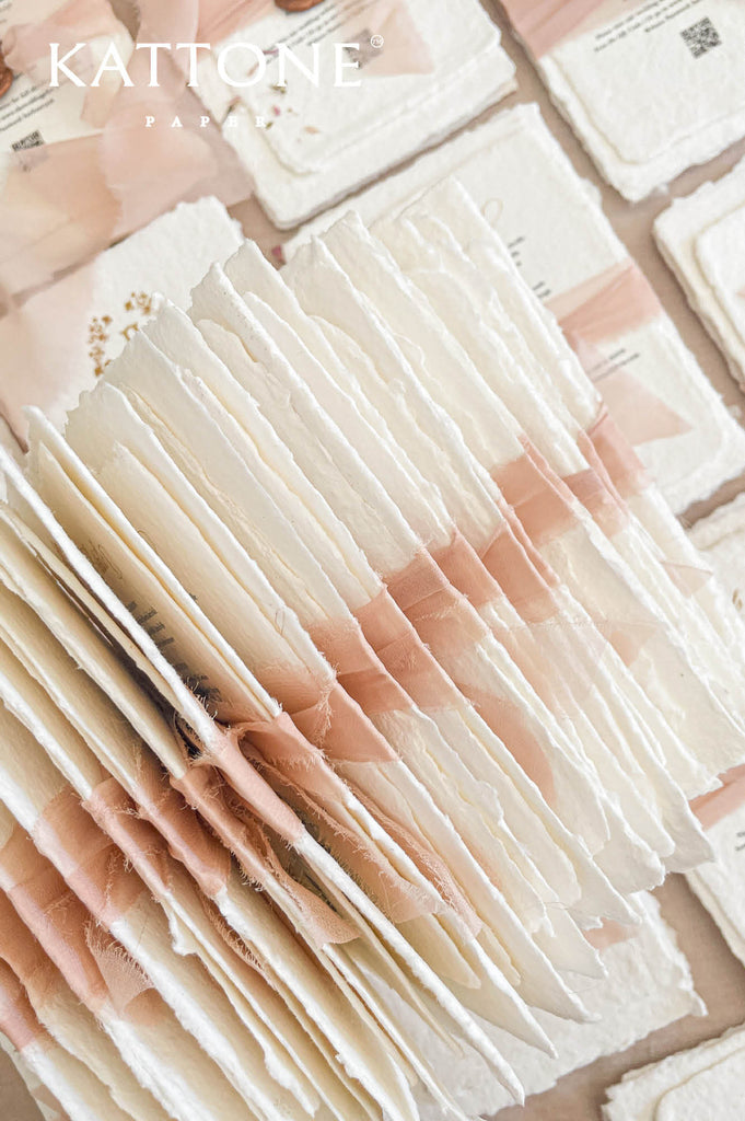 Stacked handmade paper wedding invitations and enclosure cards showcasing custom monogram crest and soft blush accents.