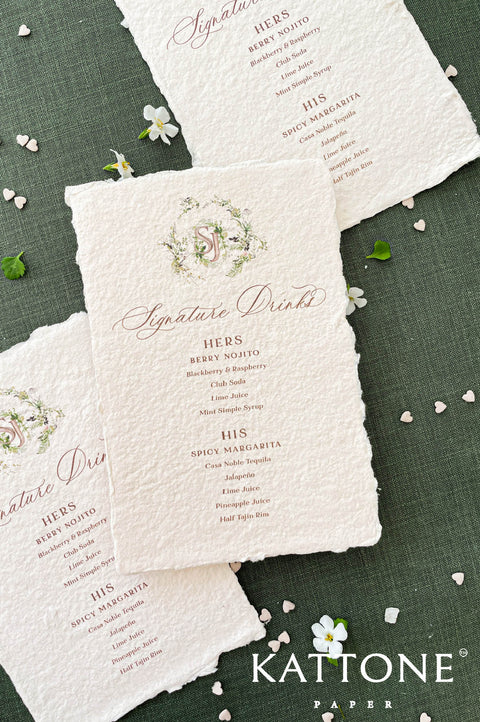 Close-up of handmade paper wedding menu featuring a custom watercolor monogram and elegant serif typography.