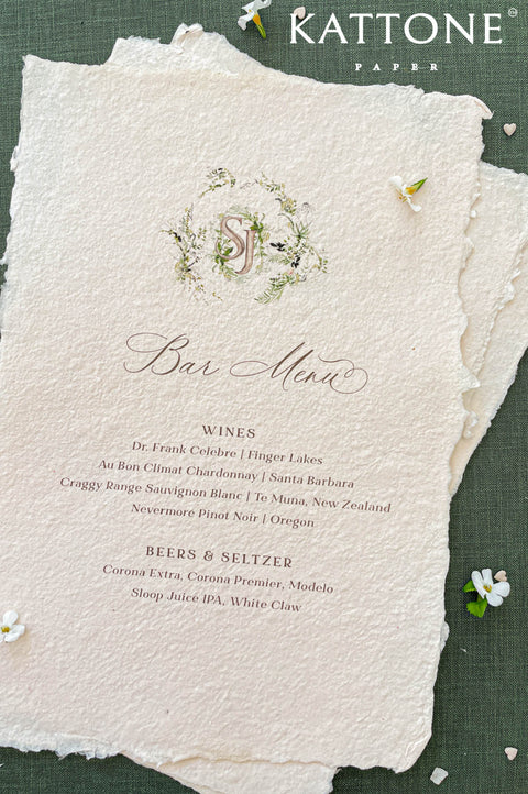 Close-up of handmade paper wedding menu featuring a custom watercolor monogram and elegant serif typography.