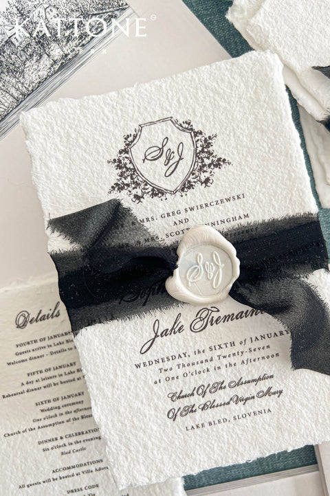 Angled overhead view of an elegant ivory handmade paper wedding invitation featuring a detailed black floral shield monogram, classic serif typography, and romantic calligraphy. The invitation is wrapped with a black sheer ribbon and finished with an ivory wax seal. A coordinating black ink illustrated envelope liner featuring a historic lakeside church and a matching wedding details card complete the European-inspired stationery arrangement.