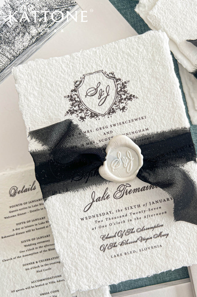 Angled overhead view of an elegant ivory handmade paper wedding invitation featuring a detailed black floral shield monogram, classic serif typography, and romantic calligraphy. The invitation is wrapped with a black sheer ribbon and finished with an ivory wax seal. A coordinating black ink illustrated envelope liner featuring a historic lakeside church and a matching wedding details card complete the European-inspired stationery arrangement.