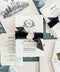 Overhead flat lay of a luxury black and white wedding invitation suite featuring ivory handmade paper invitations with personalized floral crest monograms, black sheer ribbons, and embossed ivory wax seals. Coordinating wedding details cards, ivory envelopes with black architectural illustrations of a historic lakeside church, elegant address printing, and soft blue ribbon accents are arranged on a textured blue-green background.