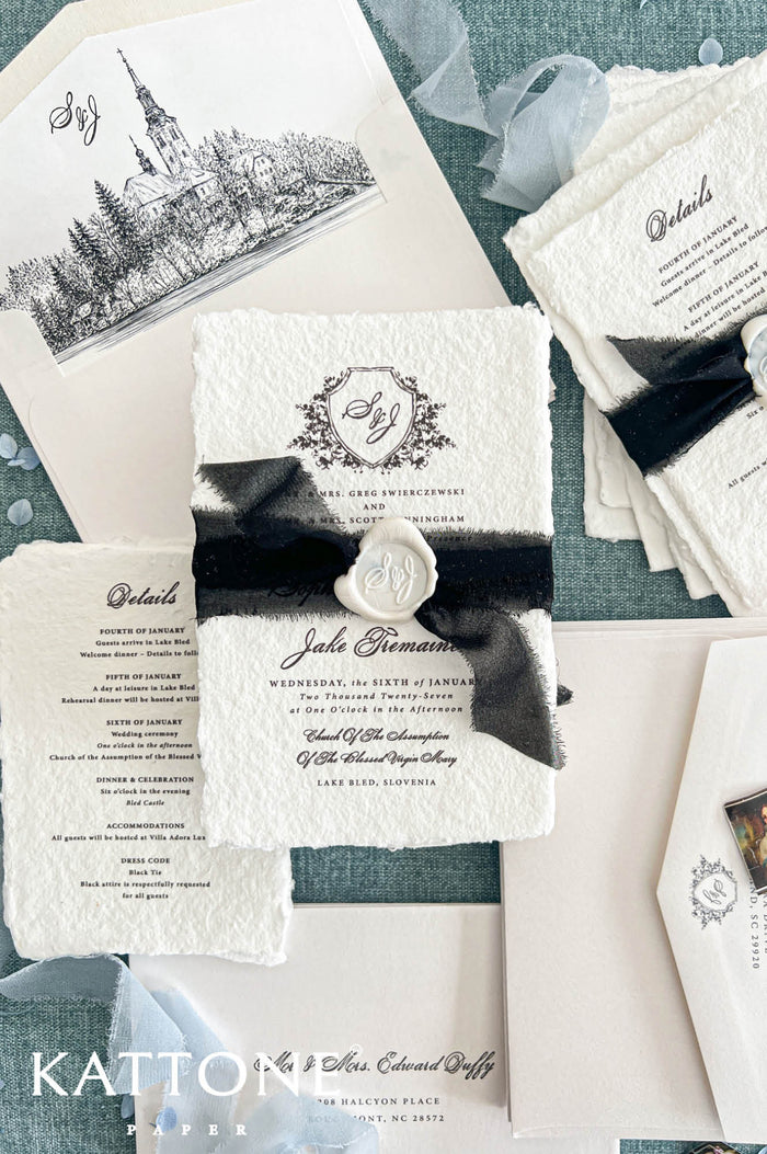 Overhead flat lay of a luxury black and white wedding invitation suite featuring ivory handmade paper invitations with personalized floral crest monograms, black sheer ribbons, and embossed ivory wax seals. Coordinating wedding details cards, ivory envelopes with black architectural illustrations of a historic lakeside church, elegant address printing, and soft blue ribbon accents are arranged on a textured blue-green background.
