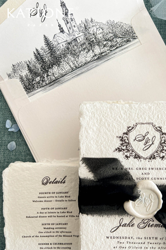 Overhead arrangement of luxury ivory wedding envelopes with custom black and white illustrated liners depicting a historic European lakeside church, woodland scenery and tranquil water. Each envelope features an elegant personalized calligraphy monogram above the detailed architectural drawing, creating a coordinated stationery design for a romantic European destination wedding.