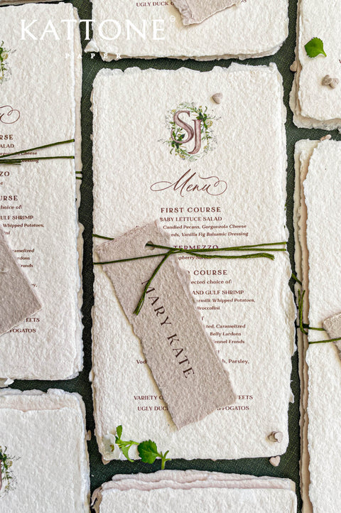 Close-up of handmade paper wedding menu featuring a custom watercolor monogram and elegant serif typography.