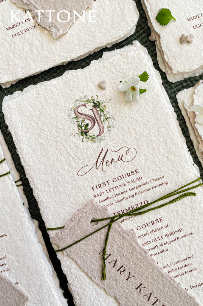 Close-up of handmade paper wedding menu featuring a custom watercolor monogram and elegant serif typography.