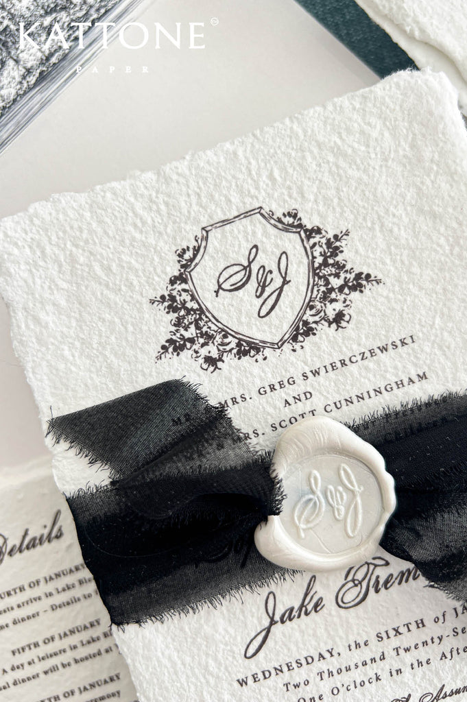Close-up photograph of a luxury ivory handmade paper wedding invitation featuring a black floral crest monogram, elegant black calligraphy, and naturally textured deckled edges. A black sheer ribbon with delicate frayed edges is tied around the invitation and secured with an embossed ivory monogram wax seal. A matching wedding details card and ivory stationery are visible in the background.