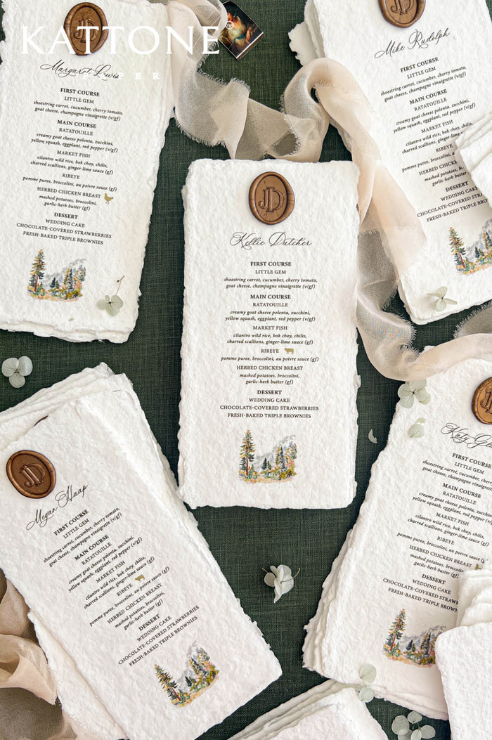 Overhead arrangement of personalized handmade wedding dinner menus with naturally deckled edges, individual guest names, antique gold monogram wax seals, and delicate watercolor pine tree illustrations, displayed with flowing ivory chiffon ribbon and scattered pale green flowers on a dark green background.