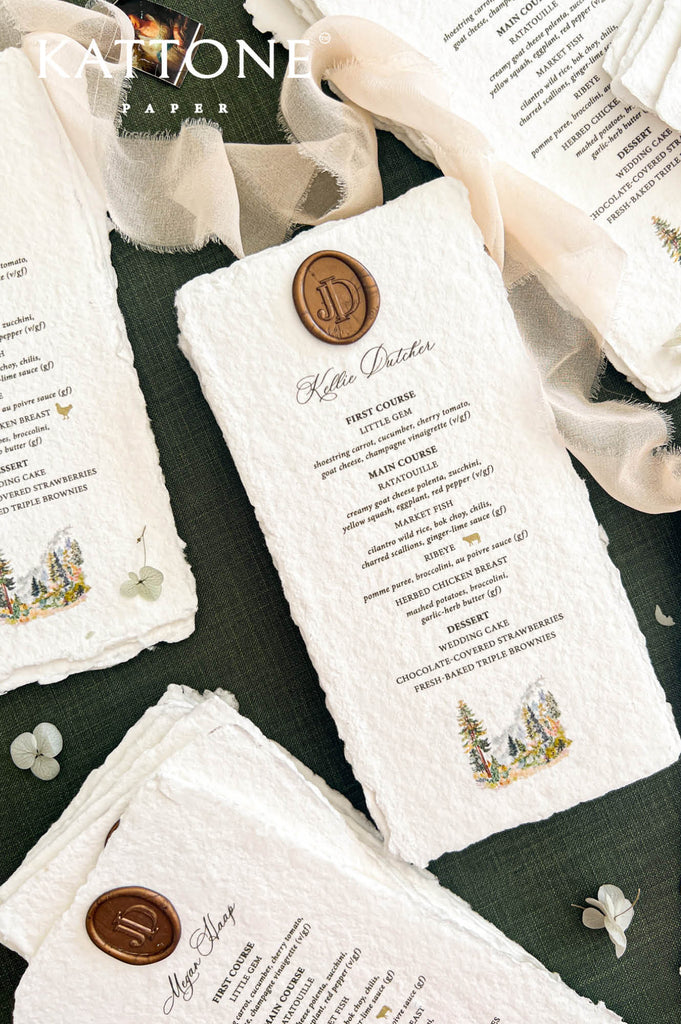 Angled overhead view of an ivory handmade paper wedding menu personalized with elegant calligraphy, a bronze monogram wax seal, custom dinner selections, and a colorful watercolor forest illustration, surrounded by matching wedding menus and flowing ivory chiffon ribbon.
