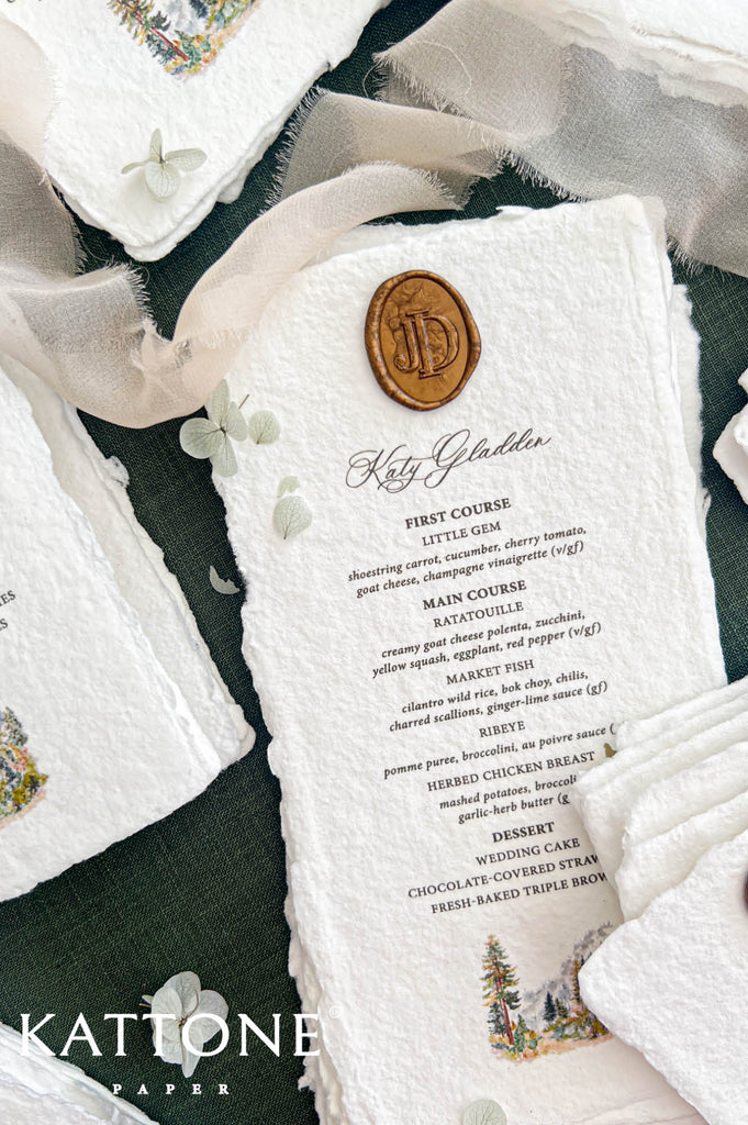 Close-up of a personalized ivory handmade paper wedding menu featuring a bronze monogram wax seal, elegant guest name calligraphy, black serif dinner menu typography, and a colorful woodland watercolor illustration at the bottom, styled with soft ivory chiffon ribbon on dark forest green fabric.