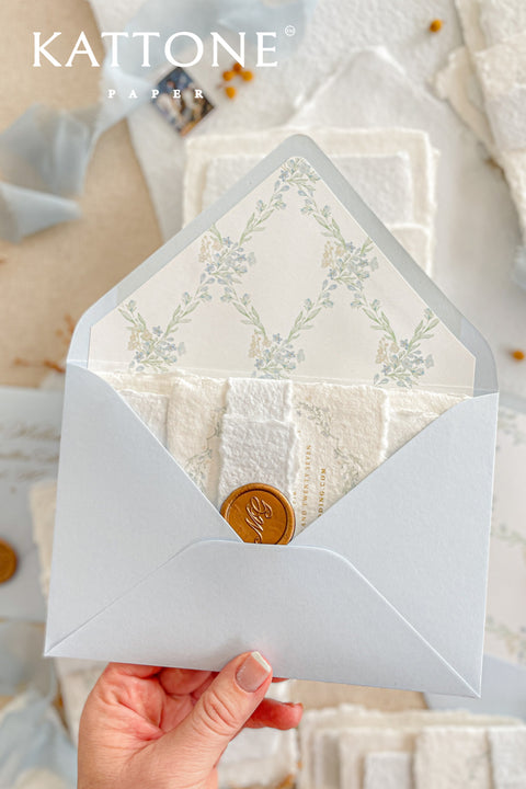 Light blue wedding envelopes with watercolor floral envelope liners held in hand, featuring soft pastel tones, wax seal detail, and fine art stationery styling.