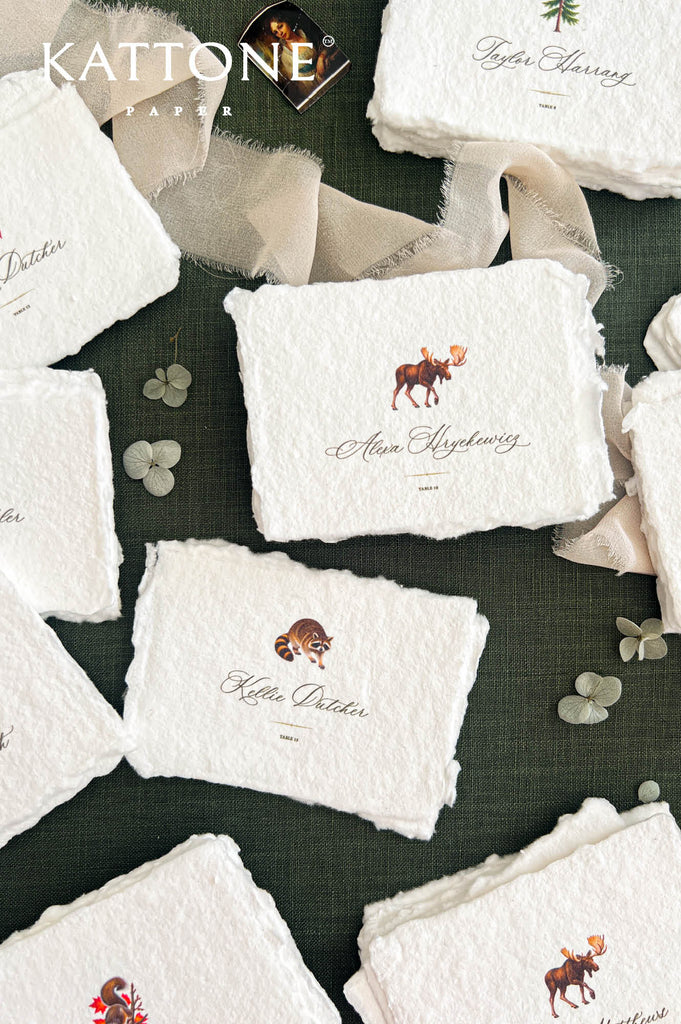 Close-up overhead view of luxury handmade paper wedding place cards featuring detailed woodland moose and raccoon illustrations above personalized calligraphy guest names and small table numbers. Thick ivory textured paper with naturally torn deckled edges, surrounded by beige ribbon and pale dried hydrangea petals on dark forest green fabric.
