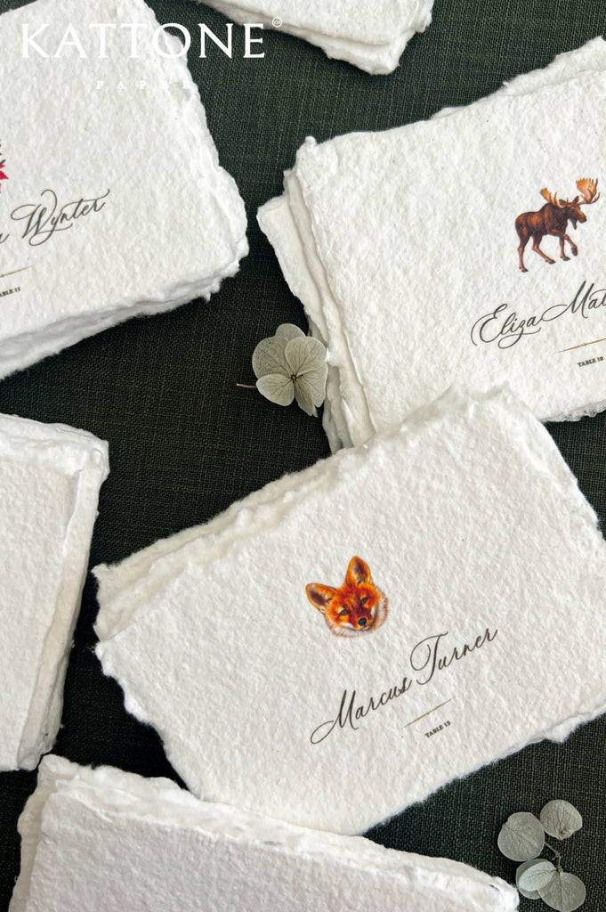 Personalized woodland wedding place cards printed on textured ivory handmade paper with natural deckled edges, featuring colorful fox and moose illustrations, elegant black guest name calligraphy, and individual table numbers. Close-up arrangement on dark forest green fabric with delicate dried flowers.