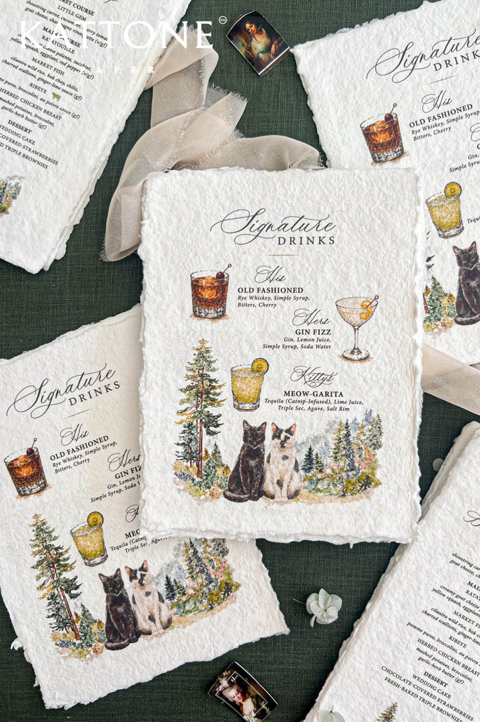 Overhead flat lay of personalized handmade paper wedding signature cocktail menus featuring illustrated drinks, a black cat and a white cat with dark markings, and watercolor forest scenery. The deckled-edge menus are arranged alongside matching wedding dinner menus, flowing ivory ribbon, and delicate flowers on a forest green background.