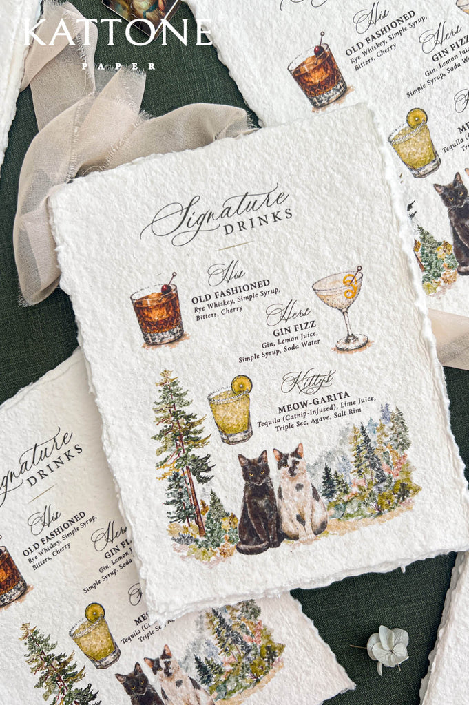 Angled close-up of an ivory handmade paper signature drinks wedding menu with elegant black calligraphy, custom cocktail names and ingredients, colorful watercolor illustrations of three cocktails, two cats sitting among pine trees, and natural deckled edges, surrounded by matching cocktail menus and soft ivory chiffon ribbon.