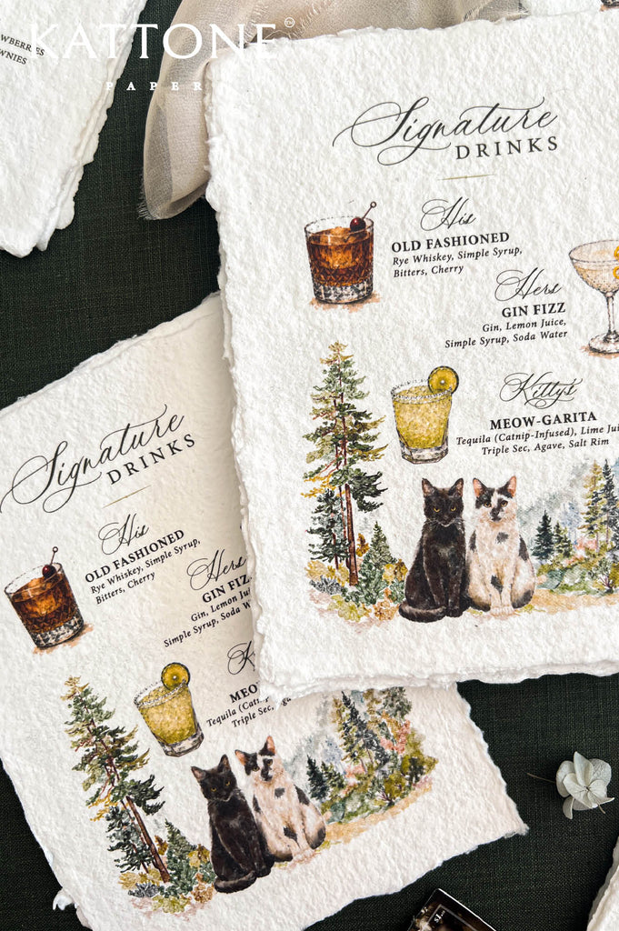 Close-up of custom signature drinks wedding menus printed on textured ivory handmade paper with deckled edges, featuring watercolor illustrations of an Old Fashioned, Gin Fizz and margarita, personalized cocktail names, two cats, and colorful woodland pine trees, styled with ivory chiffon ribbon on dark green fabric.