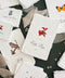 Personalized woodland wedding escort cards on textured ivory handmade paper with deckled edges, featuring colorful watercolor illustrations of a red ladybug, deer, butterfly, and evergreen tree. Individual guest names in elegant black calligraphy and table numbers are displayed against dark green fabric with flowing beige chiffon ribbon.