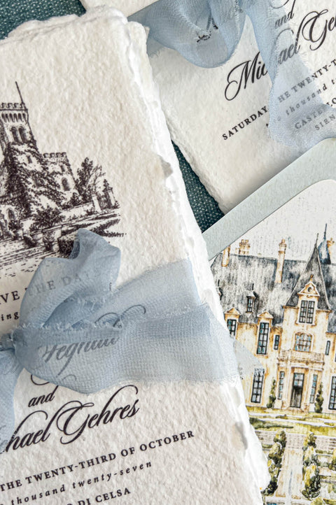 Detailed overhead close-up of ivory handmade paper Italian castle save the date cards featuring intricate black ink architectural artwork, elegant personalized calligraphy, and dusty blue chiffon bows. A coordinating pale blue wedding envelope reveals a custom watercolor château liner with formal gardens, a central fountain, and soft green foliage.