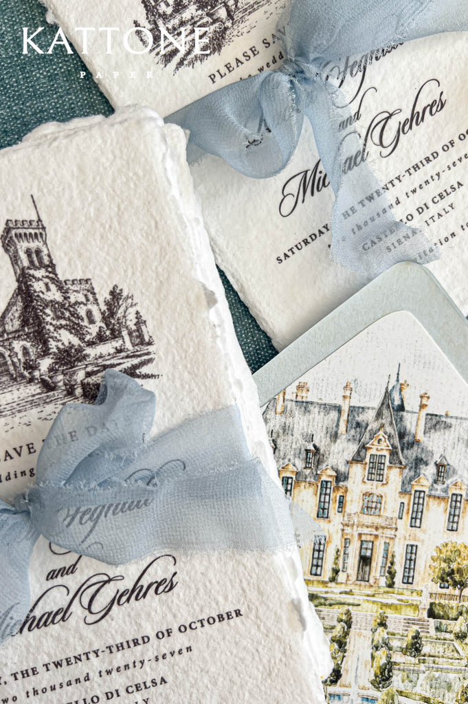 Detailed overhead close-up of ivory handmade paper Italian castle save the date cards featuring intricate black ink architectural artwork, elegant personalized calligraphy, and dusty blue chiffon bows. A coordinating pale blue wedding envelope reveals a custom watercolor château liner with formal gardens, a central fountain, and soft green foliage.