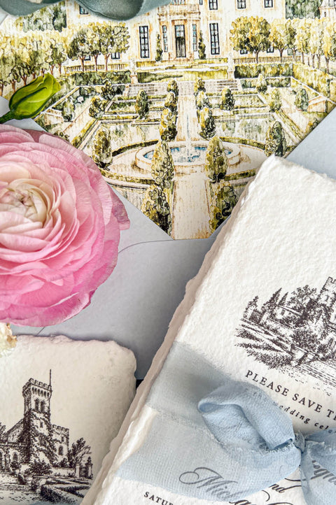 Close-up of a luxury Italian castle wedding save the date suite featuring ivory handmade paper cards with detailed black ink castle illustrations and dusty blue chiffon ribbon. A pale blue envelope reveals a custom watercolor liner depicting a grand château with symmetrical formal gardens, a central fountain, and lush greenery, styled alongside a pink ranunculus flower on textured blue linen.