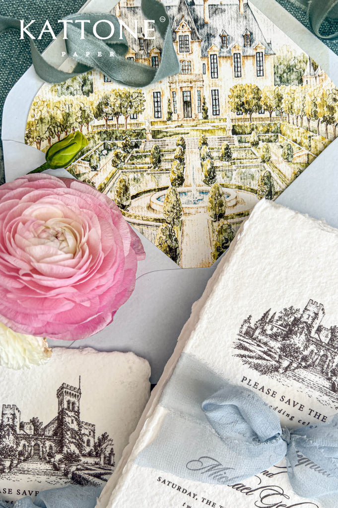 Close-up of a luxury Italian castle wedding save the date suite featuring ivory handmade paper cards with detailed black ink castle illustrations and dusty blue chiffon ribbon. A pale blue envelope reveals a custom watercolor liner depicting a grand château with symmetrical formal gardens, a central fountain, and lush greenery, styled alongside a pink ranunculus flower on textured blue linen.
