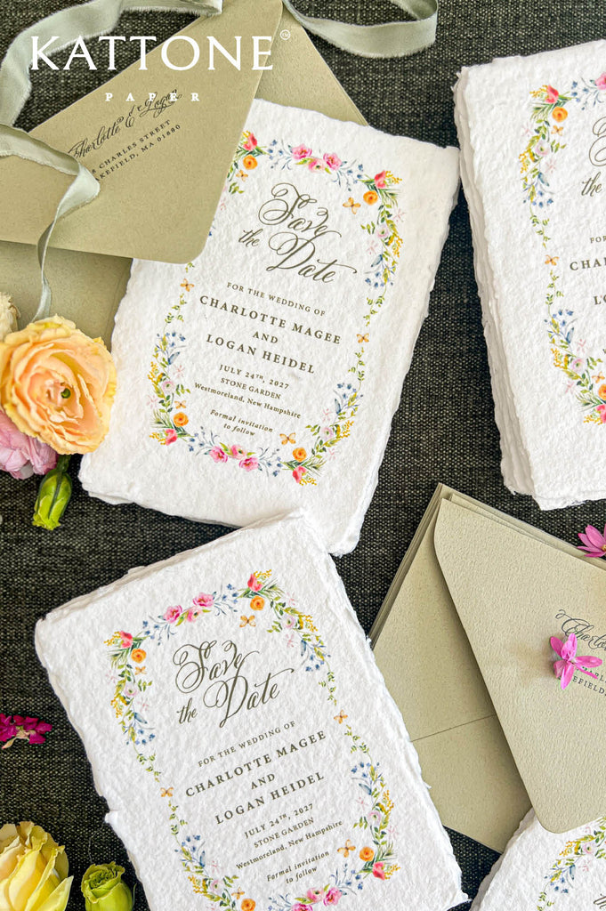 Angled flat lay of luxury watercolor floral save the date cards printed on textured ivory handmade paper with naturally deckled edges. Delicate pink, yellow, and blue flowers frame elegant calligraphy and personalized wedding details, styled with sage green envelopes, matching address printing, soft green ribbon, and fresh garden roses.