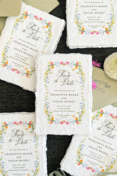 Wide overhead flat lay of romantic handmade paper save the date cards featuring colorful watercolor floral wreaths, elegant calligraphy, and personalized wedding details. Multiple ivory deckled-edge cards are arranged alongside sage green envelopes, flowing satin ribbon, pink and cream ranunculus flowers, and delicate greenery on dark textured fabric.