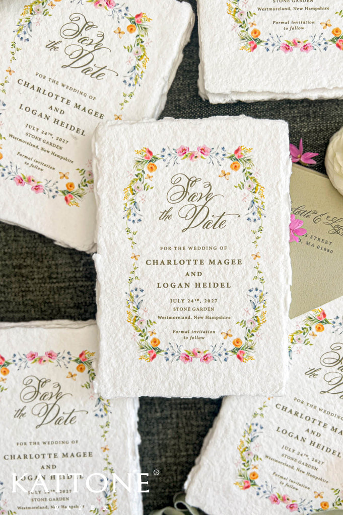 Overhead close-up of ivory handmade paper save the date cards featuring a colorful watercolor floral wreath with pink roses, peach and yellow flowers, delicate blue blossoms, and soft green foliage. Elegant calligraphy and personalized wedding details are printed inside the floral border, with multiple deckled-edge cards arranged on dark textured fabric.