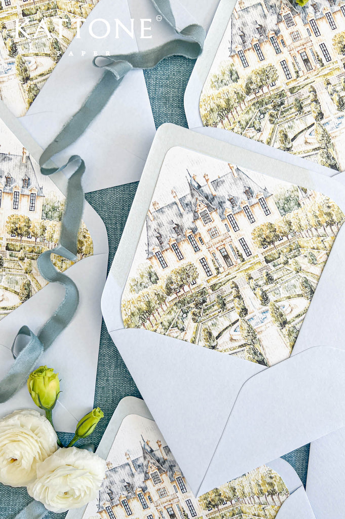 Overhead arrangement of pale blue luxury wedding envelopes with matching watercolor château envelope liners, illustrating a grand stone estate, symmetrical French gardens, fountains, and greenery. Styled with cream ranunculus, yellow rosebuds, and pink flowers.