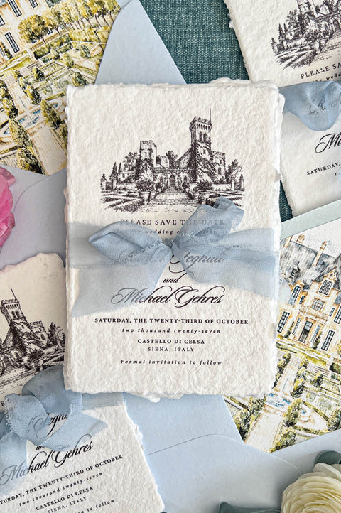 Flat lay of luxury Italian destination wedding save the date cards printed on textured ivory handmade paper with natural deckled edges and detailed black architectural castle illustrations. The cards are tied with dusty blue chiffon ribbon and accompanied by pale blue envelopes with colorful watercolor château and garden liners, surrounded by romantic flowers and decorative ribbon.