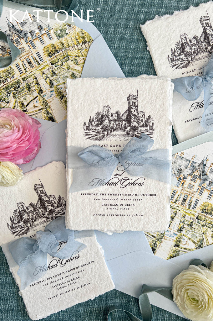 Flat lay of luxury Italian destination wedding save the date cards printed on textured ivory handmade paper with natural deckled edges and detailed black architectural castle illustrations. The cards are tied with dusty blue chiffon ribbon and accompanied by pale blue envelopes with colorful watercolor château and garden liners, surrounded by romantic flowers and decorative ribbon.