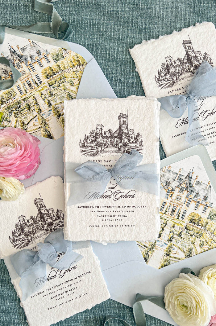 Flat lay of luxury Italian destination wedding save the date cards printed on textured ivory handmade paper with natural deckled edges and detailed black architectural castle illustrations. The cards are tied with dusty blue chiffon ribbon and accompanied by pale blue envelopes with colorful watercolor château and garden liners, surrounded by romantic flowers and decorative ribbon.