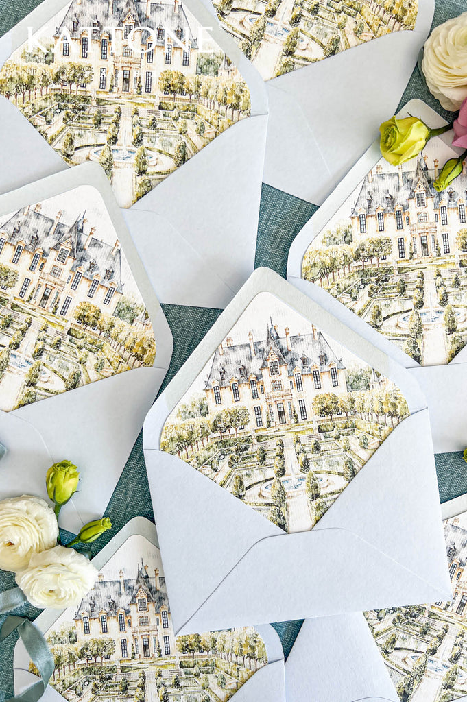Luxury powder blue wedding envelopes arranged with custom European château watercolor liners depicting formal landscaped gardens, stone pathways, and a central fountain. Styled with flowing dusty blue ribbon and ivory flowers on a blue textured surface.