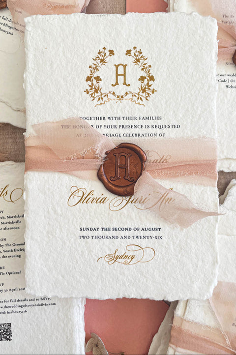 Close-up of handmade cotton paper wedding invitation featuring a gold monogram crest, textured deckle edges, and refined typography.