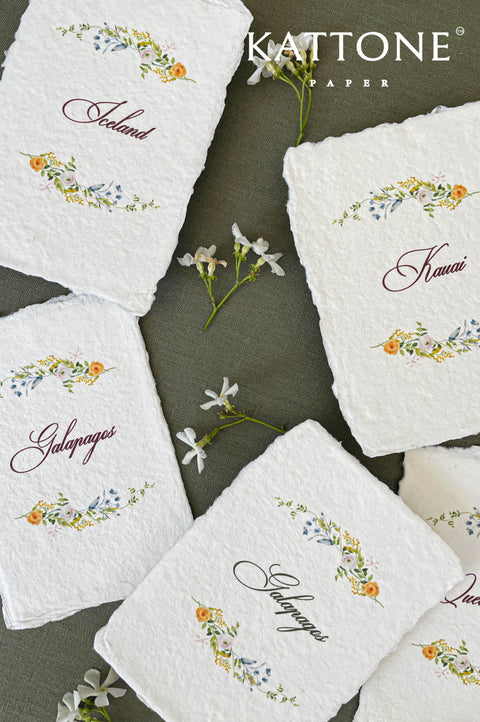 Close-up of handmade cotton paper place cards with natural deckled edges, showcasing fine watercolor floral wreath illustrations and flowing calligraphy text on textured white paper.