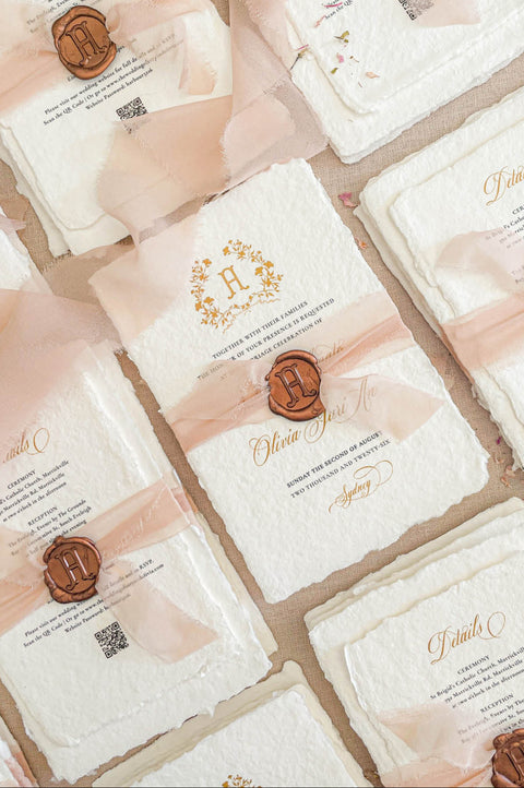 Flat lay of luxury wedding stationery suite with handmade paper invitations, details cards, monogram crest, and blush silk ribbon styling.