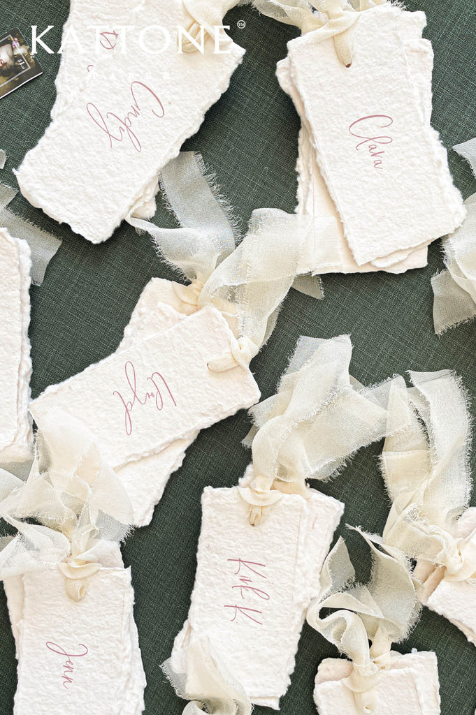 Flat lay of ivory handmade cotton paper place cards with hand-torn edges and flowing ribbon details, arranged for a fine art wedding reception table with floral accents.