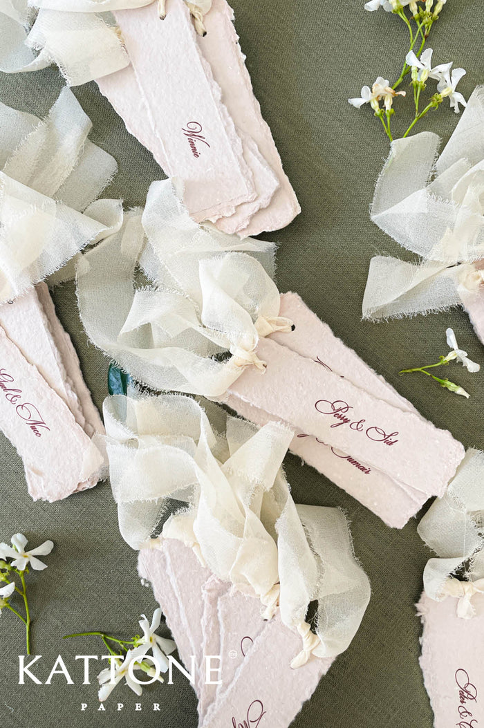 Blush handmade paper place cards with deckled edges, calligraphy names, and soft ribbon styling.
