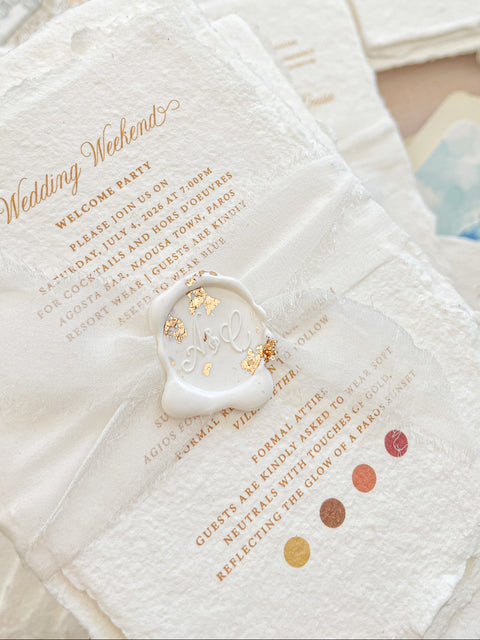 A close-up of a wedding invitation suite featuring a textured white envelope with gold foil script reading 'Wedding Weekend,' a white wax seal stamped with gold initials, and matching invitation cards in the background.