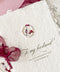 Three handmade wedding invitation cards made of textured off-white paper with rustic torn edges, decorated with small round wax seals containing dried flowers and dark red sheer ribbon bows, arranged on a pink fabric background with scattered rose petals.