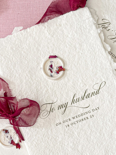 Three handmade wedding invitation cards made of textured off-white paper with rustic torn edges, decorated with small round wax seals containing dried flowers and dark red sheer ribbon bows, arranged on a pink fabric background with scattered rose petals.