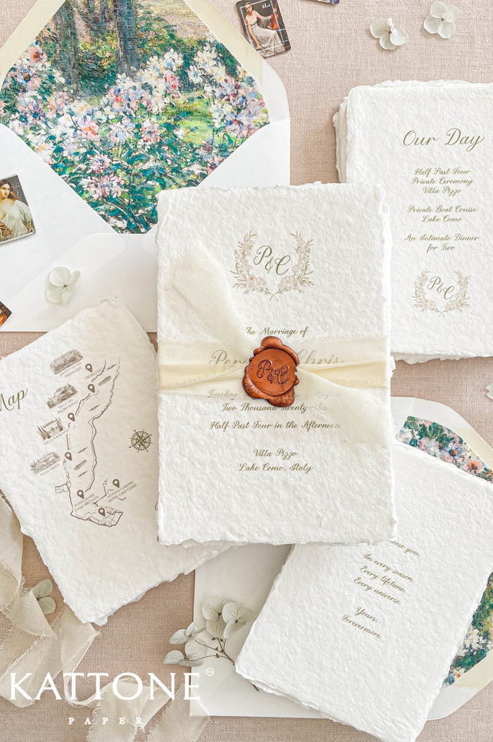 A styled flat-lay of Kattone Papers wedding stationery, featuring a central ivory textured invitation sealed with a brown wax seal, surrounded by coordinating cards, an envelope with floral artwork, and a map card on a beige fabric background.
