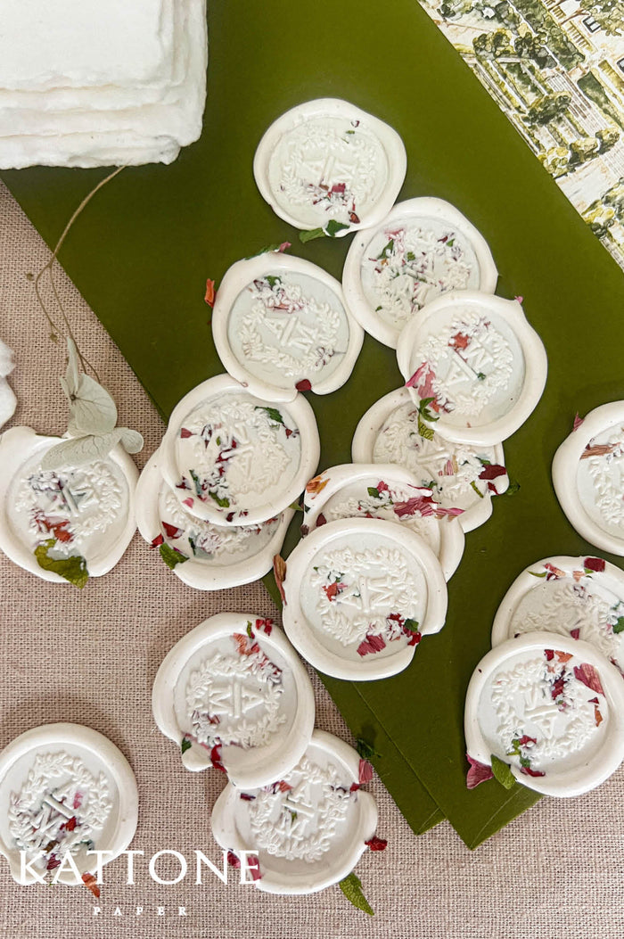 Close-up top-down view of ivory floral wedding wax seals with embossed monogram initials and embedded pink, red, and green dried flower petals, arranged on olive green paper and neutral linen