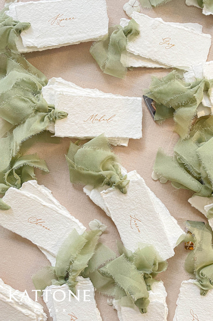 Top-down flat lay of personalized handmade paper wedding place cards with textured deckled edges, warm calligraphy guest names, and long sage green frayed silk ribbon ties, styled with dried flowers on a neutral linen background